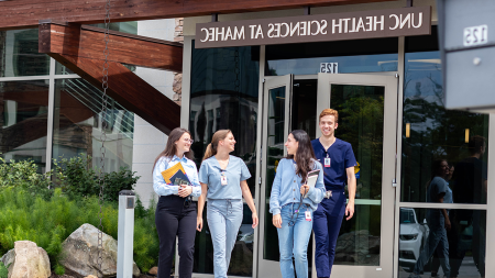 Four medical students wearing scrubs and holding books exit building at the School of Medicine's Asheville campus. Sign above front door reads 