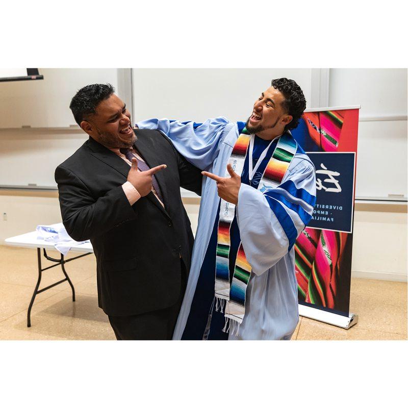 Student in graduation gown and Exitos sash smiles with staff member